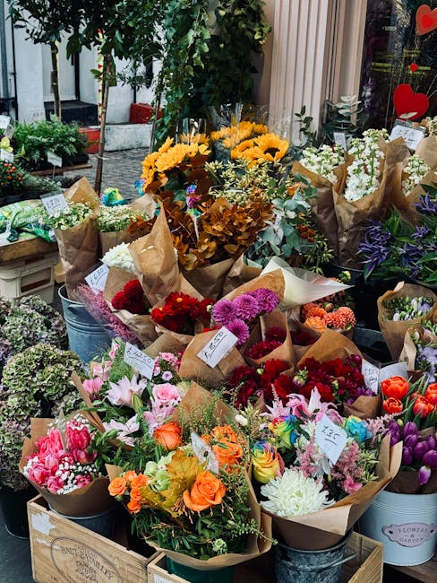 A display of fresh flower bouquets arranged outdoors in buckets and boxes at a flower shop, featuring a variety of vibrant blooms including sunflowers, roses, lilies, tulips, and chrysanthemums in sha