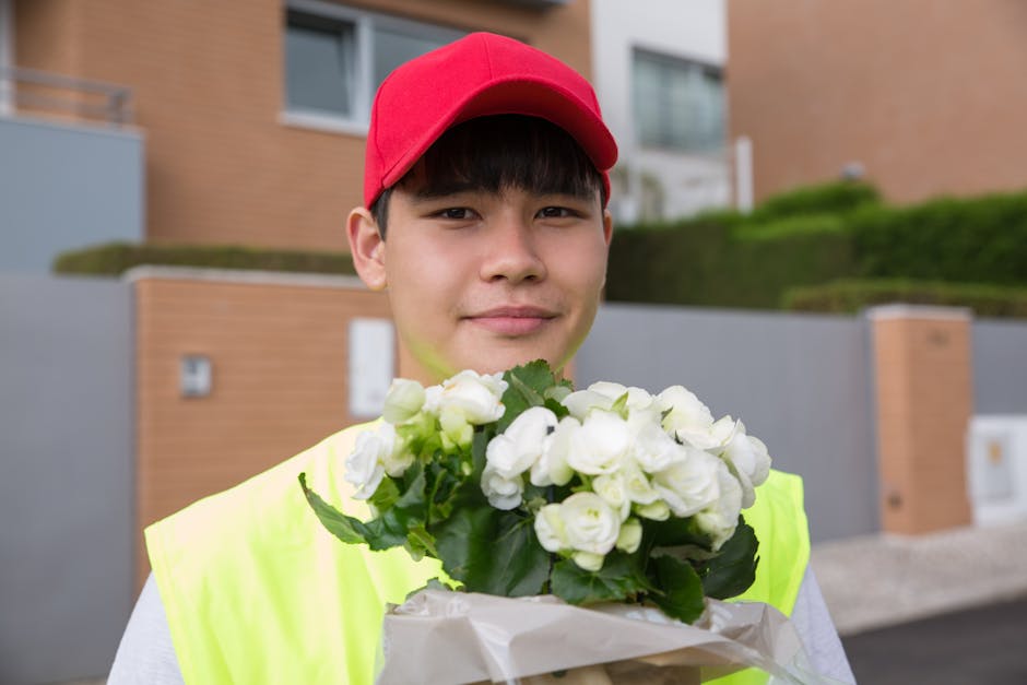 A young male flower delivery courier wearing a bright yellow safety vest and a red cap, holding a bouquet of white gardenias wrapped in brown paper, standing outdoors in front of modern residential bu