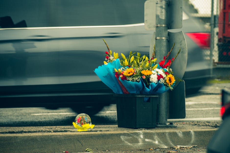 A vibrant bouquet of fresh flowers sits in a black box on the ground at a street corner, partly wrapped in bright blue paper with yellow, red, white, and orange blooms including sunflowers, lilies, an