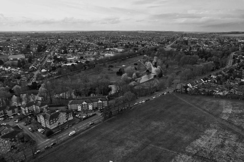 A black-and-white aerial photograph of Cowley, featuring a large park with trees and a pond near the center, surrounded by residential houses, apartment buildings, and a busy road with cars. The scene