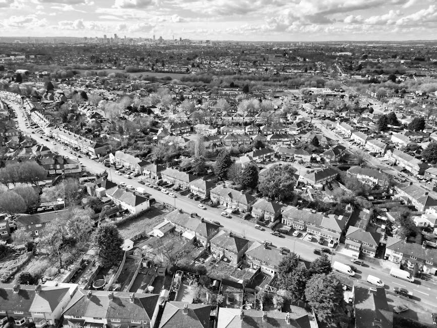 A black-and-white aerial photograph of a suburban residential area showing a street lined with numerous detached and semi-detached houses, each with front gardens and driveways. The houses have pitche