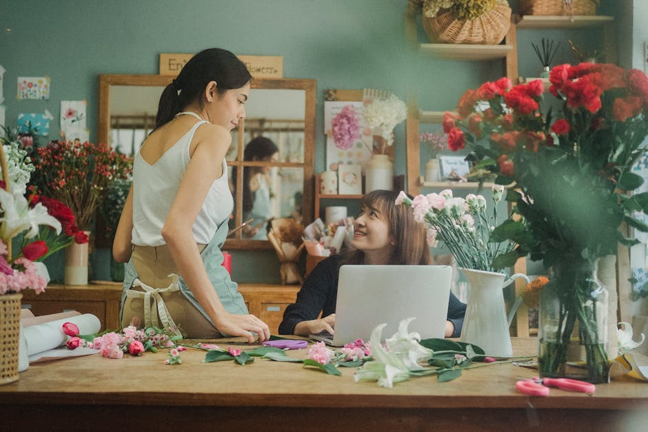 A florist and a customer engage in a conversation inside a flower shop. The florist, a woman with dark hair tied back, wears a white sleeveless top and a beige apron, standing beside a wooden table sc